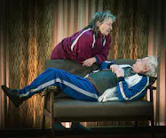 Per Sandberg och Gunilla Röör, iklädda träningsoveraller, i rollerna som Rut och Ragnar. Per ligger på en soffa och Gunilla står lutad över honom. 