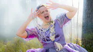 Affisch till föreställningen Den skrattande drottningen. Skådespelaren Karin Paulin i lila klänning, flera lager pärlhalsband. Trolsk, disig skog i bakgrunden. 
