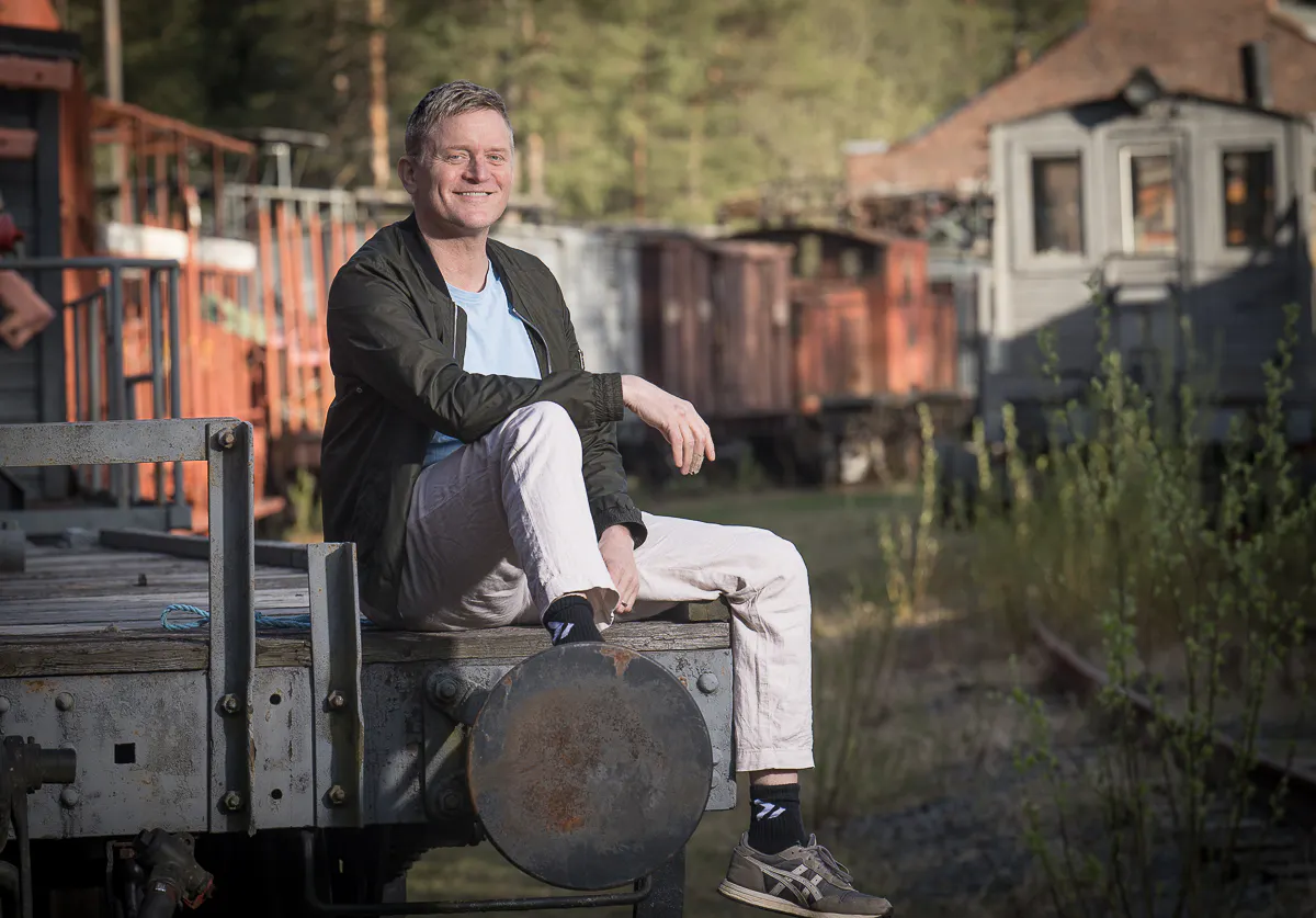 Martin Sundbom, regissör, Kabaré Solstugan. Sitter på en sliten tågvagn, tågvagnar på räls i bakgrunden. 